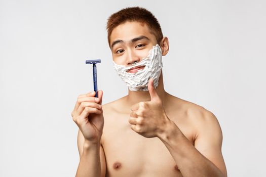 Beauty, people and hygiene concept. Pleased good-looking asian man recommend razor or shaving cream, show thumb-up and smiling pleased while face in cream, standing white background.