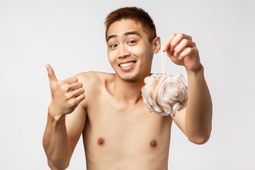 Beauty, people and hygiene concept. Portrait of satisfied cute asian man with naked torso standing white background, showing bath sponge and thumb-up, recommend good product for showering.