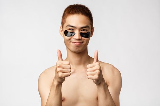 Beauty, people and lifestyle concept. Portrait of satisfied, carefree handsome asian man with naked torso, looking pleased, smiling show thumb-up recommend eye-patched to look fit and healthy.