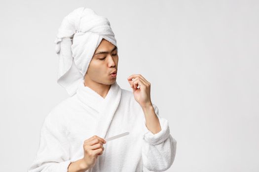 Beauty, spa and leisure concept. Portrait of handsome young man have day-off, enjoying quarantine, wear bathrobe and bath towel on head, polishing nails, standing white background.