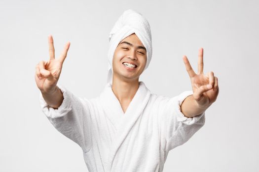 Beauty, spa and leisure concept. Upbeat handsome asian man in bathrobe and bath towel, show peace signs and smiling with rejoice, express enthusiasm, stand white background.