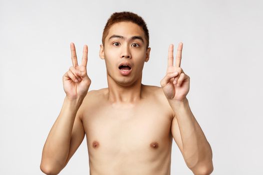 Beauty, people and home concept. Shocked young asian man with naked torso, showing peace sign or quotation marks, gasping startled, hear strange news, express disbelief, white background.