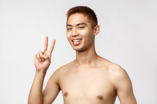 Beauty, people and leisure concept. Handsome enthusiastic asian man with naked torso, show peace sign and smiling pleased, apply skincare cosmetics cream under eye and stand white background.