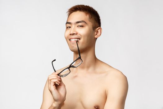 Beauty, people and lifestyle concept. Close-up portrait of confident and sassy asian man with naked torso, biting rim of glasses and smiling pleased, looking camera know something interesting.