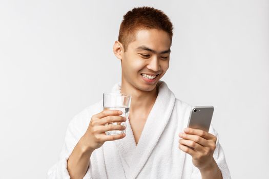 Beauty, spa and technology concept. Handsome happy asian man on vacation, checking news in mobile phone internet network, hold smartphone and drink water from glass, smiling. Copy space