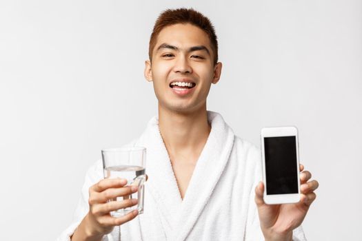 Beauty, spa and technology concept. Portrait of enthusiastic asian man in bathrobe showing his healthcare application, smartphone screen with water consumption tracker, hold glass and smiling.