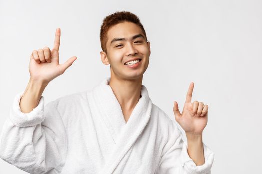 Beauty, spa and leisure concept. Portrait of cheerful, handsome enthusiastic asian man in bathrobe, pointing fingers up, showing way and dancing pleased, smiling joyfully, white background.