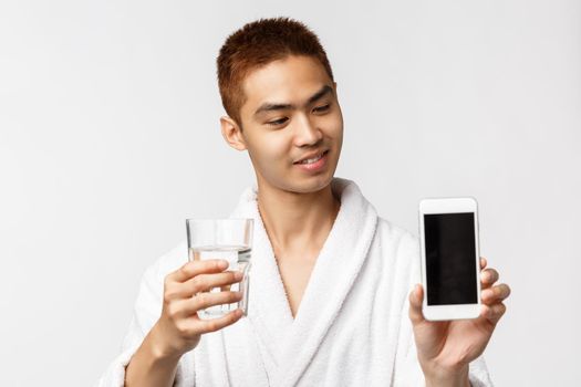 Beauty, spa and technology concept. Pleased handsome asian man in bathrobe, showing smartphone display, drinking water from glass and looking at mobile phone app, white background.