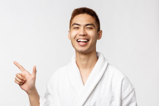 Beauty, spa and leisure concept. Portrait of cheerful asian man in bathrobe, enjoying vacation, pointing finger left, showing way, laughing and smiling pleased, standing white background.