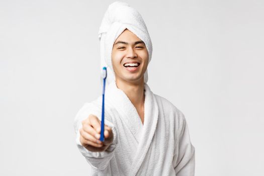 Beauty, spa and leisure concept. Young happy asian man in bathrobe and bath towel, showing toothbrush and smiling cheerful, taking care of personal hygiene, standing white background.