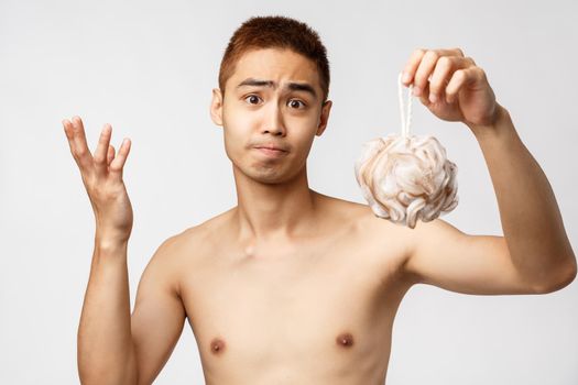 Beauty, people and hygiene concept. Confused and sad handsome asian man with bare torso, standing naked holding sponge and shrugging puzzled, look perplexed camera, white background.