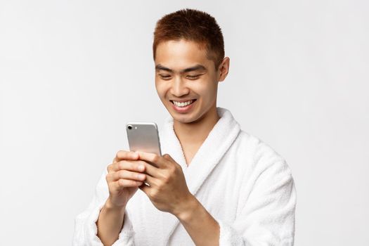 Beauty, spa and technology concept. Handsome asian man in bathrobe looking pleased at smartphone display, texting friends from vacation hotel resort, standing white background with mobile phone.