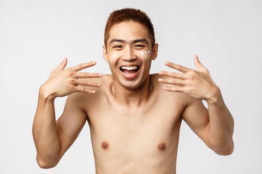 Beauty, people and leisure concept. Close-up portrait of enthusiastic, amused asian handsome man with bare torso, shaking hands and laughing, smiling upbeat, apply facial cream. Copy space