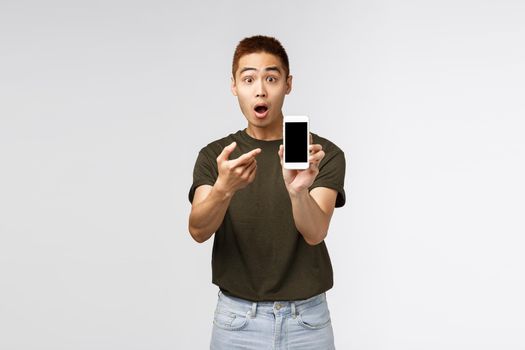 Technology, online lifestyle and communication concept. Surprised and amazed asian guy shocked pointing at mobile phone display to show something stunning and amazing, grey background.