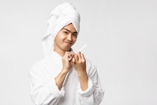 Beauty, spa and leisure concept. Portrait of silly good-looking asian man in bathrobe and bath towel, smiling pleased, polishing nails and make manicure, standing white background.