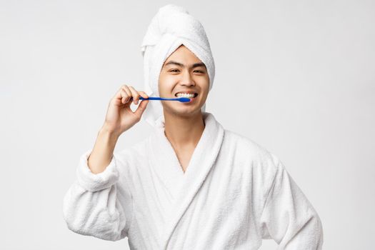 Beauty, spa and leisure concept. Upbeat happy asian man in bathrobe and bath towel, brushing his teeth with toothbrush and smiling, taking care of personal hygiene, white background.