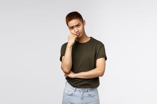 Portrait of gloomy, sad asian guy in green t-shirt, pouting and looking camera upset, lean on palm, feeling uneasy about missing good party, envy people outside while sitting on quarantine.