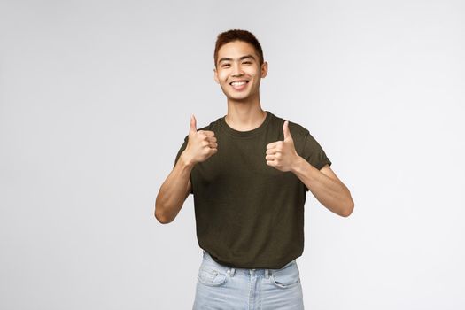 Cheerful asian man, male student recommend online learning courses, tutoring foreign language during quarantine codi19, show satisfied thumb-up and smiling pleased, grey background.