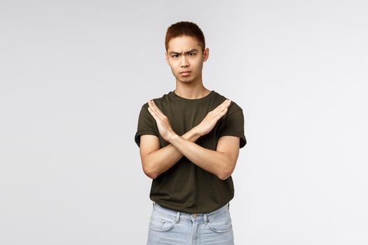 Serious-looking angry asian man make cross sign and frowning disappointed, saying no, disapprove and disagree, give negative feedback, prohibit and forbid, grey background. Copy space