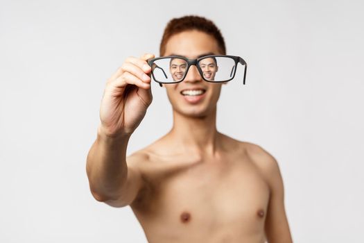 Beauty, people and lifestyle concept. Close-up portrait of enthusiastic asian man with naked torso, showing glasses, looking through eyewear and smiling upbeat, standing white background.