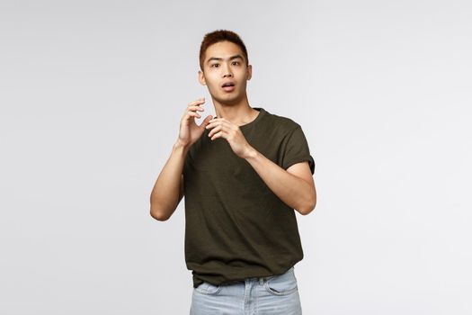 Portrait of startled young asian guy, male student gasping and jumping scared as someone appeared unexpected, raising hands up defense, stare terrified at camera, standing grey background.