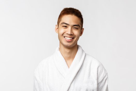 Beauty, spa and leisure concept. Young smiling, cheerful asian man grinning at camera delighted after awesome massage at hot springs summer hotel resort, wear bathrobe, white background.