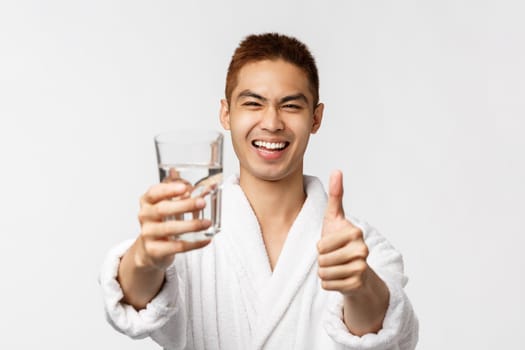 Beauty, spa and technology concept. Portrait of satisfied, happy laughing asian man in bathrobe, show thumbs-up and glass with water, smiling pleased, recommend be healthy, white background.