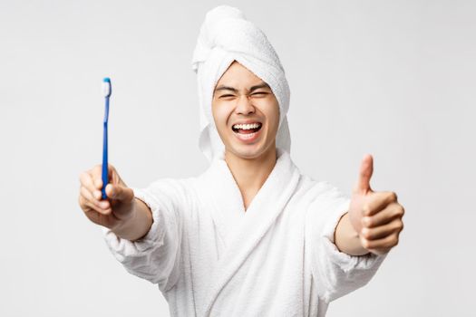 Beauty, spa and leisure concept. Portrait of enthusiastic handsome asian man in bathrobe and bath towel, show toothbrush with satisfied face, make thumb-up in appoval, recommend healthcare product.