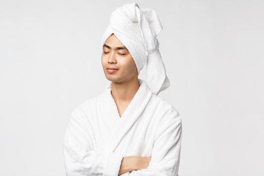 Beauty, spa and leisure concept. Portrait of relaxed, calm and peaceful happy asian man enjoying vacation, close eyes and smiling, wear bathrobe and towel over head, white background.