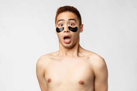 Beauty, people and lifestyle concept. Portrait of surprised, startled young asian man with naked torso and black eye-patches, gasping amazed, open mouth and stare impressed camera, white background.