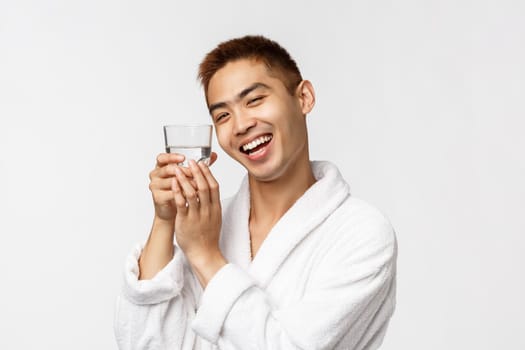 Beauty, spa and technology concept. Portrait of cute and silly asian man play around, laughing and smiling as hugging glass of water, wear bathrobe at home during self-distancing, white background.