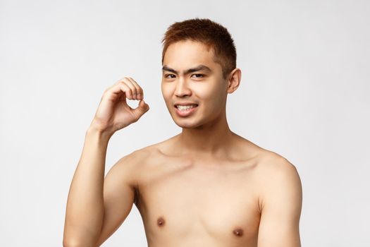 Beauty, people and home concept. Portrait of funny handsome asian man with naked torso, pointing behind or shaking hand, grimacing look unsure, standing white background. Copy space