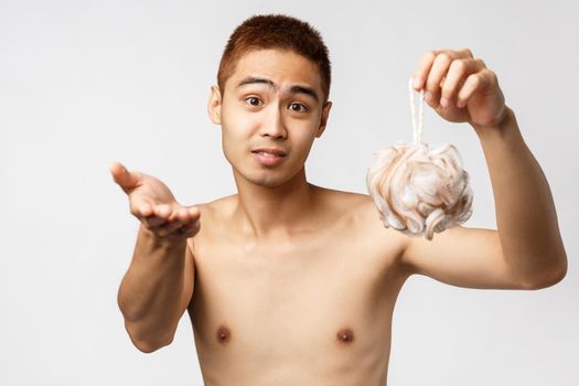 Beauty, people and hygiene concept. Portrait of handsome young asian man holding bath sponge and pointing hand at camera, asking to wash his back, standing white background.