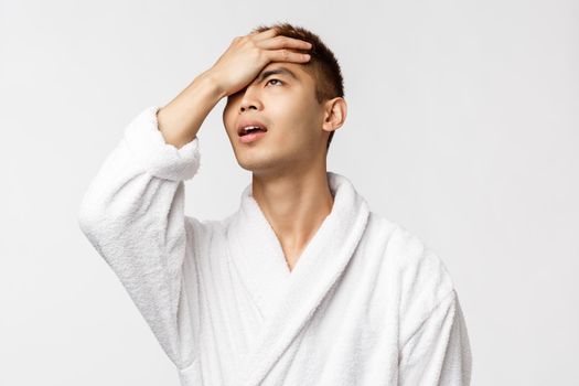 Beauty, spa and leisure concept. Portrait of upset, sighing and concerned asian man, facepalm looking up regret something, feel failure and frustration, wear bathrobe, white background.