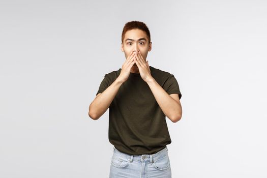 Surprised and amused asian man gasping, cover mouth and look camera impressed, express astonishment and happiness for friend, hear great news, standing grey background pleased.