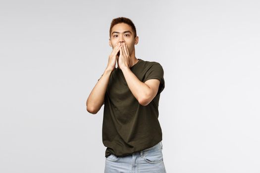 Portrait of shocked, speechless asian guy gasping, cover mouth and express shook and surprised with eyes, gazing camera at unbeliavable amazing thing, standing grey background.