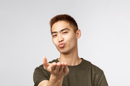 People, different expressions and lifestyle concept. Close-up portrait of coquettish, cheeky asian romantic guy, sending air kiss at camera, folding lips and blow mwah with hand, grey background.