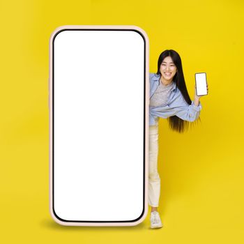 Cute asian young girl excited peeking out big giant vertical cell phone holding smaller one in hand with white blank screen isolated on yellow background. Great offer. Product placement. Copy space.