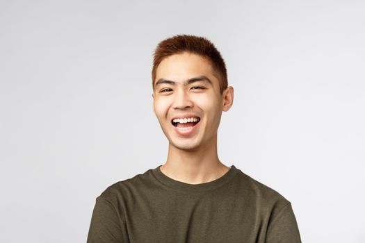 Close-up potrait of enthusiastic handsome young hipster guy, asian male student laughing, talking to friends videochatting, smiling pleased, feel rejoice and excitement, stand grey background.