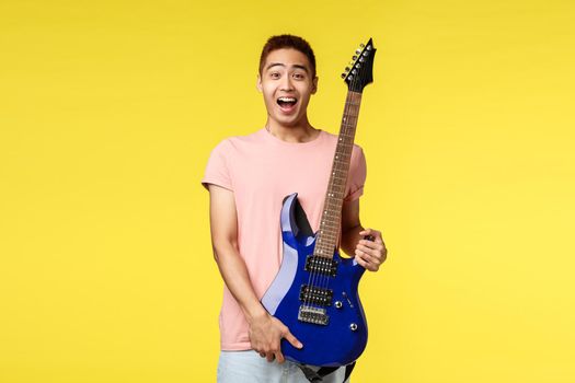 Lifestyle, leisure and youth concept. Portrait of handsome cheerful asian man in pink t-shirt, laughing and smiling excited, hold blue electric guitar and look surprise, receive gift new instrument.