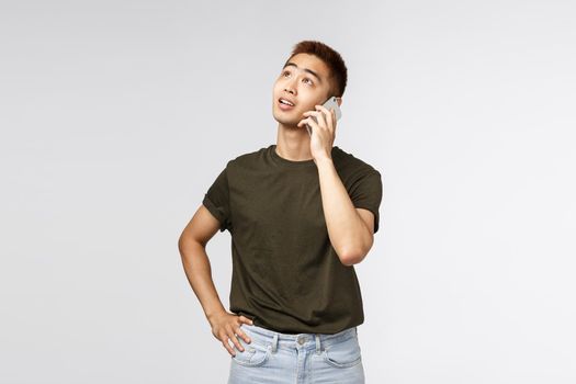 Technology, online lifestyle and communication concept. Thoughtful good-looking asian male talking on mobile phone, look dreamy up, thinking what answer, order food delivery, grey background.