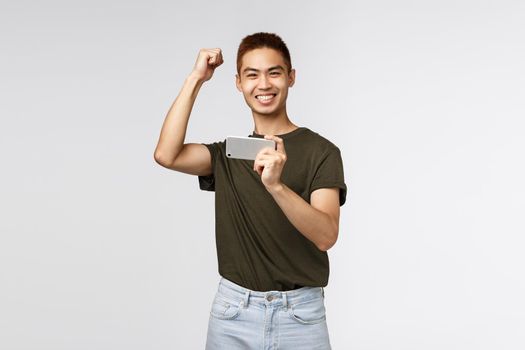 Technology, online lifestyle and communication concept. Portrait of rejoicing, happy asian man raising hand up in yes, hooray emotion, winning, hold mobile phone, pass level in smartphone game.