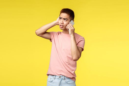 Technology, communication and lifestyle concept. Portrait of troubled, indecisive young asian guy look puzzled up, talking on mobile phone, having tough conversation, yellow background.