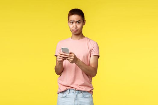 Technology, communication and lifestyle concept. Portrait of alarmed asian guy in pink t-shirt, using mobile phone text alarmed at bad news he read internet, standing bothered and frustrated.