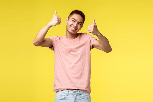 Lifestyle, travel and people concept. Happy asian man in pink t-shirt satisfied with awesome product, show thumbs-up and smiling broadly, guarantee you like it, approve promo, yellow background.