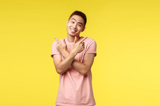 Silly and cute asian man in pink t-shirt smiling sween and caring, tilt head lovely as pointing sideways to both good variants, pointing left and right, showing two choices, stand yellow background.