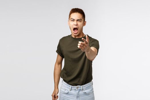 Portrait of angry heartbroken asian boyfriend being cheated on, accuse person for betraying him, pointing fingers and cursing, shouting and blaming someone, grey background.