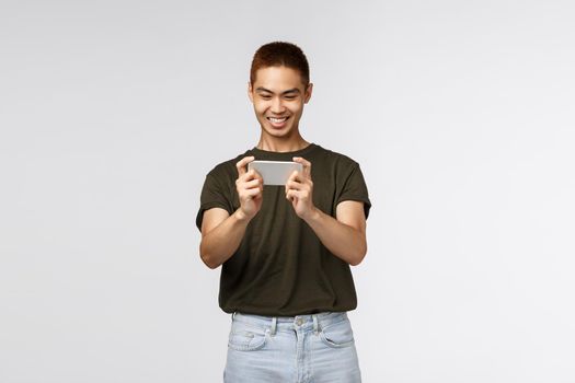 Technology, online lifestyle and communication concept. Happy pleased young asian male using mobile phone, smiling cheerful as playing new smartphone app, grin satisfied, grey background.