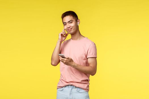 Technology, communication and lifestyle concept. Cheerful asian man in pink t-shirt, using mobile phone, put on wireless earphones to contact friend, listening music, stand yellow background.