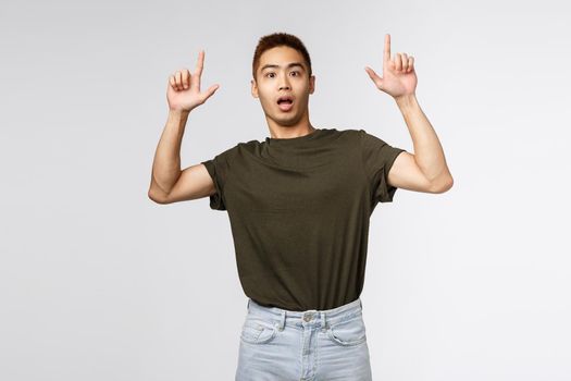 Portrait of shocked and nervous asian man look at camera surprised, pointing fingers up, showing something strange and concerning, asking question about item, grey background.